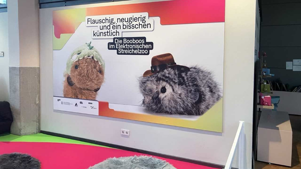 Zwei plüschige Hamster mit Hüten auf einem bunten Poster im Ausstellungsraum.